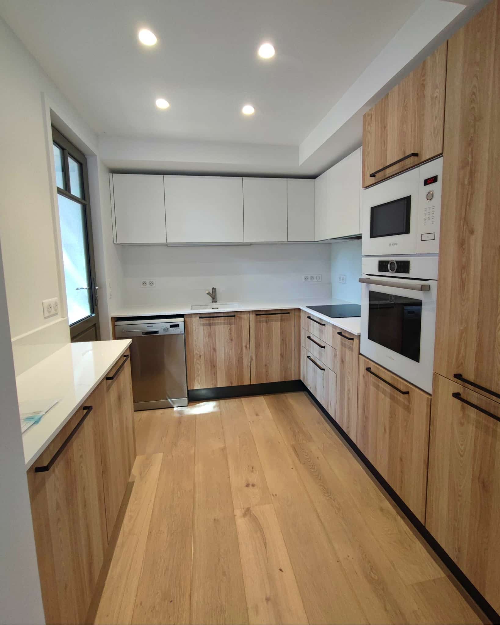 Cuisine moderne bois et blanc avec électroménager intégré Cuisine moderne en L avec meubles bas en bois clair, placards hauts blancs, plan de travail blanc et électroménager Bosch/Siemens intégré. Sol parquet.