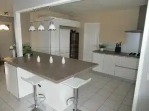 Cuisine moderne ouverte avec îlot central en bois clair, tabourets blancs, et luminaires suspendus. Meubles bas blancs, plaque de cuisson, hotte.