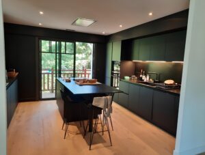 Cuisine moderne minimaliste noire avec îlot central, tabourets et plancher bois clair. Grande baie vitrée sur balcon verdoyant, spots et éclairage LED.