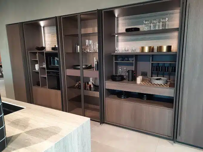 Armoire de cuisine moderne en bois foncé avec portes coulissantes ouvertes, four intégré, étagères et vaisselle organisée.