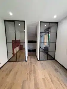 Couloir lumineux avec sol en bois, portes coulissantes vitrées noires menant à une chambre et salle de bain.