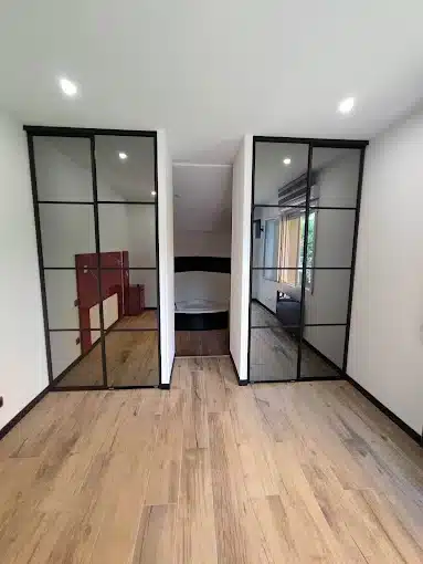 Couloir lumineux avec sol en bois, portes coulissantes vitrées noires menant à une chambre et salle de bain.