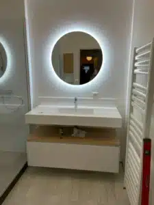 Salle de bain moderne: miroir LED rond lumineux au-dessus d'un meuble vasque flottant, douche vitrée et sèche-serviettes blanc.