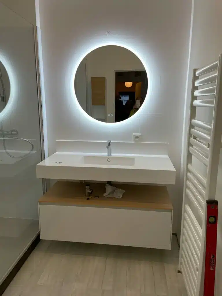 Salle de bain moderne: miroir LED rond lumineux au-dessus d'un meuble vasque flottant, douche vitrée et sèche-serviettes blanc.
