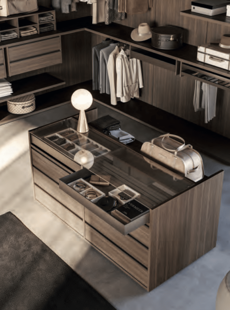 Dressing Luxueux en Bois Sombre avec Îlot de Rangement Dressing luxueux en bois sombre avec îlot central vitré, vêtements suspendus et lampe globe design.